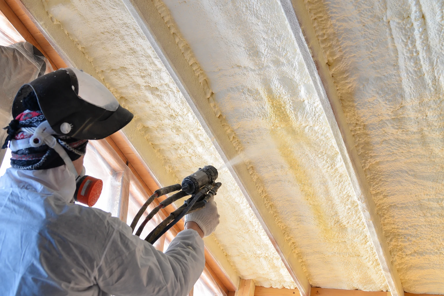A worker in protective gear sprays expanding foam insulation between roof rafters.