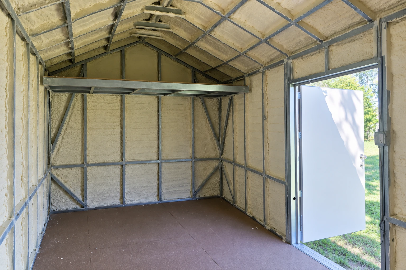 A metal-framed outbuilding is insulated with pale spray foam across its walls and pitched ceiling, beside an open door.