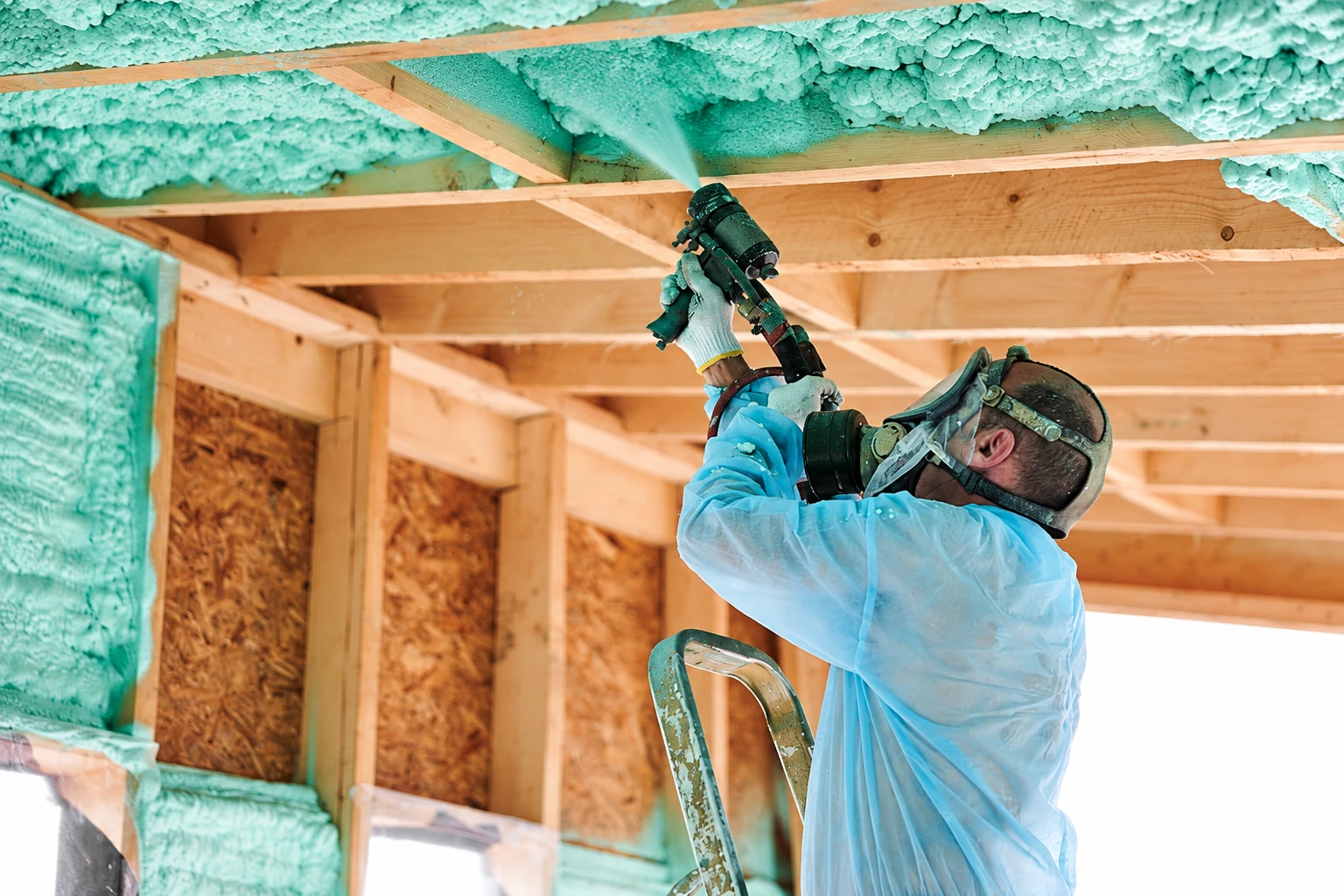 Worker in protective coveralls and a respirator sprays green foam insulation onto the underside of a wooden ceiling.