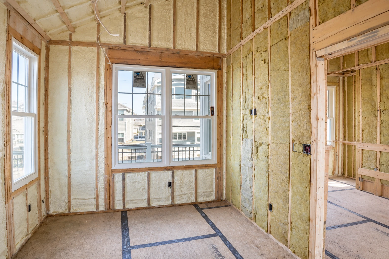 A framed room has pale spray foam insulation filling wall and ceiling cavities around large windows.